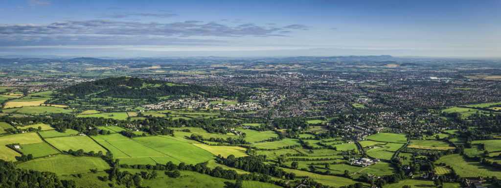 Aerial panoramic vista over the green patchwork landscape of the Cotswold escarpment, pasture and farms, to the City of Gloucester, the Severn Vale and Welsh mountains beyond. ProPhoto RGB profile for maximum color fidelity and gamut.